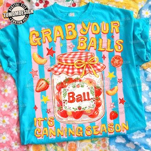 May include: A bright blue t-shirt featuring a graphic of a glass canning jar filled with strawberries. The text reads "GRAB YOUR BALLS" and "IT'S CANNING SEASON". The jar has a red and white checkered lid and is surrounded by stars and fruit.