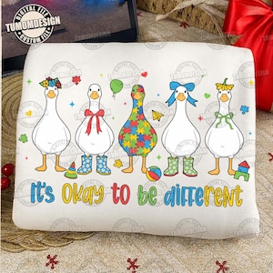 Peut inclure: Graphique blanc avec cinq oies de dessin animé, chacune ornée d'accessoires différents tels que des nœuds, des fleurs et des motifs de puzzle. Le texte "It's okay to be different" est affiché en dessous.