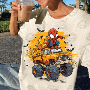 May include: Beige t-shirt featuring a Halloween-themed graphic. The design includes a cartoon Spider-Man character sitting on an orange monster truck loaded with carved pumpkins. Bats and spiderwebs complete the festive scene.