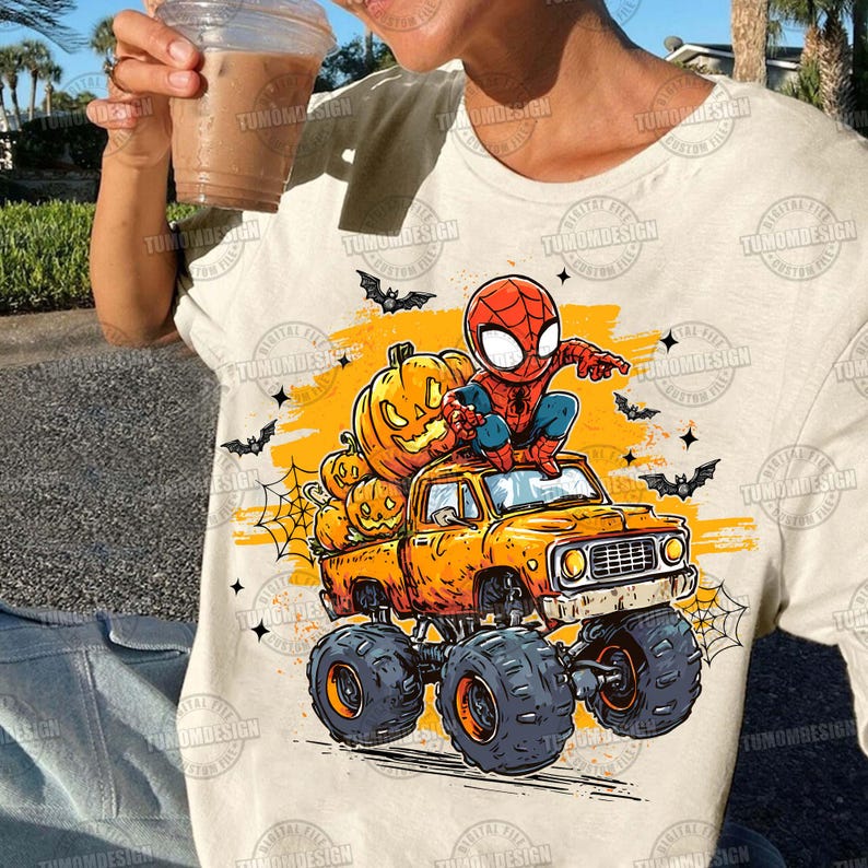 May include: Beige t-shirt featuring a Halloween-themed graphic. The design includes a cartoon Spider-Man character sitting on an orange monster truck loaded with carved pumpkins. Bats and spiderwebs complete the festive scene.