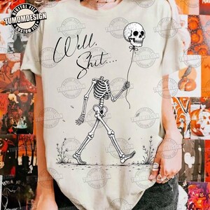 May include: A cream-colored t-shirt featuring a black line art illustration of a skeleton walking and holding a skull-shaped balloon. The text "Well, Shit...." is printed in a cursive script above the skeleton, which is surrounded by small floral sketches.