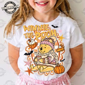 May include: A white t-shirt featuring a graphic of Winnie the Pooh wearing a pink knit hat and sweater, holding a mug. The design includes text reading WINNIE THE POOH, pumpkins, a slice of pie, bats, and stars in orange and pink tones.