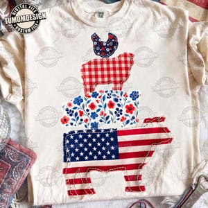 May include: Cream-colored t-shirt featuring a stacked animal design. The bottom layer is a cow with an American flag pattern, followed by a pig with a floral design, a sheep with a red and white gingham pattern, and a chicken with a blue floral pattern.
