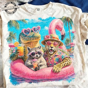 May include: A cream-colored t-shirt featuring a vibrant illustration of a crocodile, raccoon, and leopard wearing sunglasses and tropical shirts, floating on a pink inflatable ring with a flamingo. The image has a tropical theme.