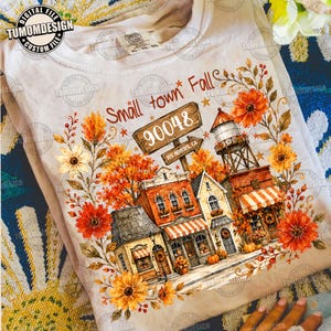 May include: A light-colored t-shirt featuring a graphic of a small town street scene during autumn, surrounded by orange and yellow flowers. The text reads Small town Fall, 90048, and Los Angeles, CA. The design includes a water tower and buildings.