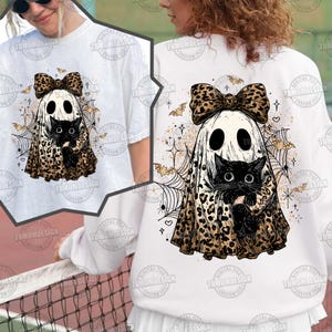 May include: White t-shirt and sweatshirt featuring a Halloween ghost design. The ghost wears a leopard print bow and cloak, holding a black cat. Spiderwebs, bats, and stars complete the spooky design.