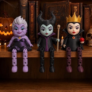 Peut inclure: Trois figurines de personnages stylisées assises sur une étagère. La figurine de gauche est violette avec des cheveux blancs et une robe noire. Celle du centre est noire avec des cornes et un bâton. Celle de droite porte une couronne dorée.