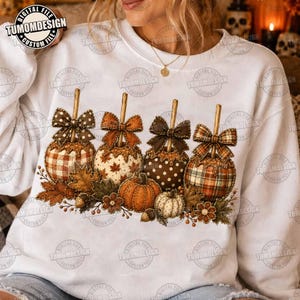 May include: A white long-sleeve sweatshirt featuring a graphic print of four decorated pumpkins with bows, autumn leaves, and acorns. The pumpkins display various patterns including plaid, polka dots, and maple leaves in shades of brown and orange.