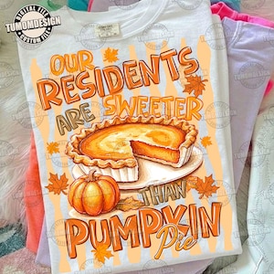 Może przedstawiać: Biały t-shirt z grafiką przedstawiającą ciasto dyniowe na talerzu oraz całą dynię. Tekst brzmi: Our residents are sweeter than pumpkin pie. Projekt zawiera pomarańczowe napisy oraz rozrzucone pomarańczowe liście klonu na jasnopomarańczowym tle.