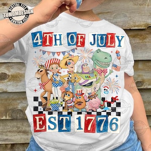Puede incluir: Camiseta blanca con un gráfico colorido de personajes animados, fuegos artificiales y el texto "4th of July EST 1776". El diseño incluye elementos patrióticos rojos, blancos y azules, perfecto para las celebraciones del Día de la Independencia.