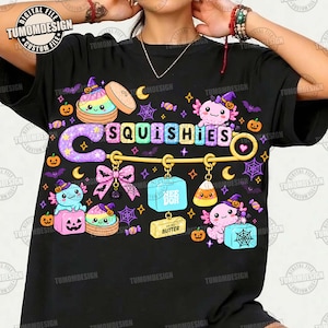 May include: A black t-shirt featuring a large graphic of a gold safety pin with the text SQUISHIES. The design includes colorful axolotls, pumpkins, candy, spiderwebs, bats, a pink bow, a butter block, and a blue cube labeled NEE DOH.