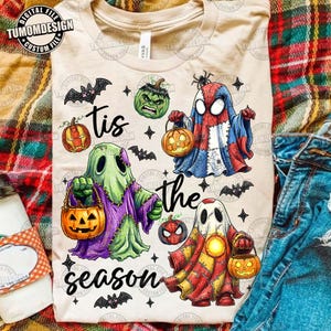 May include: A beige t-shirt featuring a graphic print of three ghost-like figures dressed as superheroes, each holding a jack-o'-lantern. The text 'tis the season' is printed in black script. Additional graphics include small bats, stars, and pumpkins.