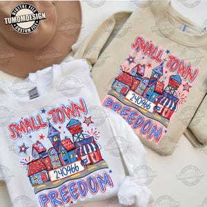 Könnte beinhalten: Zwei Sweatshirts, eines in Weiß und eines in Beige, mit einer farbenfrohen Grafik. Das Design zeigt eine Stadtansicht mit den Worten "SMALL TOWN FREEDOM" und der Nummer "240966". Die Grafik verwendet die Farben Rot, Weiß und Blau.
