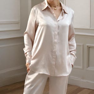 Può includere: Un completo pigiama in seta rosa chiaro. Il set comprende una camicia abbottonata con colletto e maniche lunghe e pantaloni abbinati. Il tessuto ha una lucentezza sottile e l'outfit è indossato da una persona.