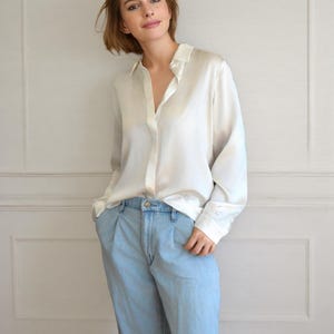 以下が含まれることがあります： 白いシルクのボタンダウンシャツと水色のデニムジーンズを着た女性。シャツは長袖で、クラシックな襟が付いています。ジーンズはハイウエストでリラックスしたフィット感です。シンプルでエレガントな服装です。