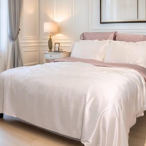 May include: A bed featuring a smooth, off-white duvet cover and matching pillowcases, layered with a dusty rose-coloured flat sheet folded over the top edge. The bedding set is displayed on a bed frame in a room with white panelled walls and light wood flooring.