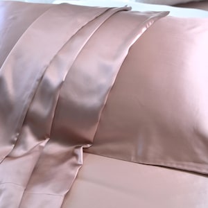 Peut inclure: Une taie d'oreiller en satin rose poudré avec une texture douce et soyeuse. La taie d'oreiller est pliée en deux et a un léger éclat.