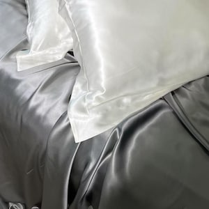 May include: A bed with white pillowcases and a grey satin sheet. The pillowcases are smooth, contrasting with the slightly creased satin sheet. The bedding has a luxurious appearance, with a soft sheen and elegant drape.