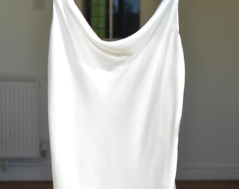 Puede incluir: Un vestido de satén blanco con cuello de pico y tirantes finos. El vestido está colgado en una percha negra.