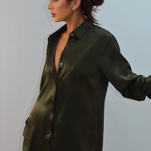 May include: Dark olive green button-down shirt with long sleeves. The shirt has a silky, satin-like sheen and is partially unbuttoned. The model is wearing black pants.