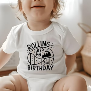May include: A white baby grow with black text that reads "Rolling into my 3rd birthday". The text is surrounded by a black and white illustration of a rollercoaster with a car at the top of the hill.