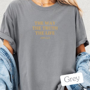 Op de afbeelding: Een grijs T-shirt met de tekst "THE WAY THE TRUTH THE LIFE JOHN 14:6" in goud gedrukt.