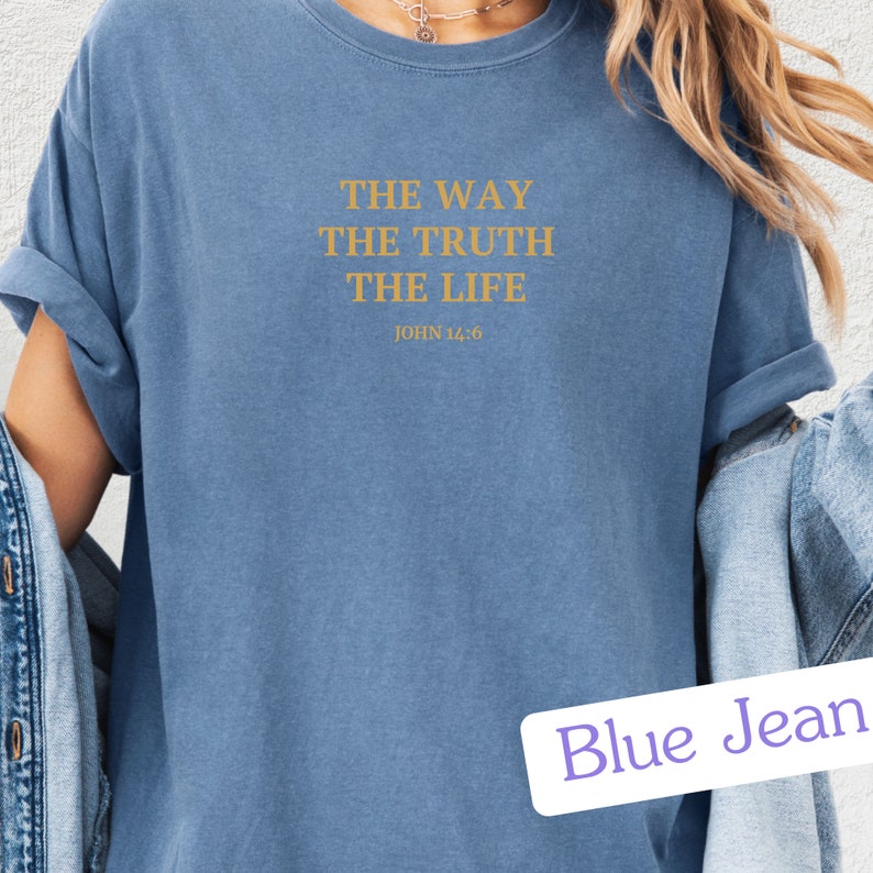 Op de afbeelding: Een blauw T-shirt met de tekst "THE WAY THE TRUTH THE LIFE JOHN 14:6" in goud gedrukt op de voorkant. Het T-shirt wordt gedragen door een persoon met lang bruin haar en een spijkerjack.