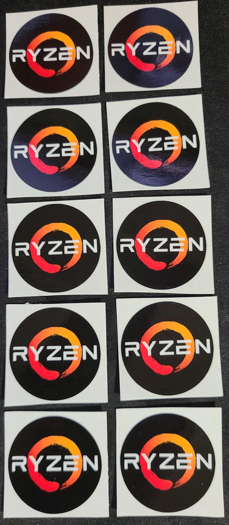 May include: Ten white square stickers with a black circle in the center. The circle has a red and orange brushstroke design with the word "RYZEN" in white letters.