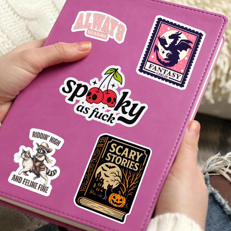 May include: A pink journal covered in stickers. Stickers include the text "ALWAYS READING", "Spooky as fuck", a cherry design, a stamp with a dragon, and a sticker with a cat riding a raccoon. The sticker says "RIDDIN' HIGH AND FELINE FINE".