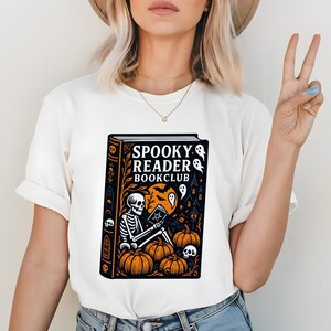 Pode incluir: T-shirt branca com um design de Halloween. O design inclui um esqueleto lendo um livro com o texto "Spooky Reader Bookclub", ab&oacute;boras, fantasmas e uma casa assombrada. A obra de arte &eacute; em preto, laranja e branco.