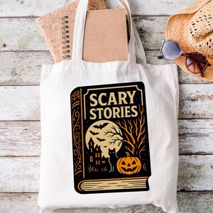May include: A white canvas tote bag featuring a black and orange graphic of a book titled "Scary Stories." The design includes a full moon, bats, a haunted house, a black cat, and a jack-o'-lantern. A notebook and sunglasses are visible.