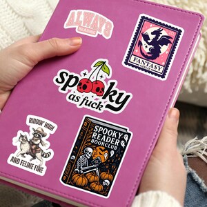 Pode incluir: Um di&aacute;rio rosa adornado com v&aacute;rios autocolantes. Os autocolantes exibem frases como "Always Reading", "Spooky as Fuck" e "Fantasy Girlie". Outros autocolantes apresentam um par de cerejas, um gato num guaxinim e um esqueleto a ler um livro.