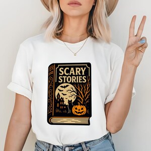 May include: White t-shirt featuring a Halloween-themed graphic. The design includes a book with the words "SCARY STORIES" in a vintage font, a full moon, bats, a haunted house, a black cat, and a jack-o'-lantern.