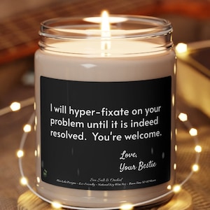 May include: A brown glass candle with a black label that reads "I will hyper-fixate on your problem until it is indeed resolved. You're welcome. Love, Your Bestie". The candle is lit and the flame is visible.