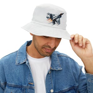 Peut inclure: Un chapeau bob blanc avec un motif d'avion de chasse en vol. L'avion est noir et gris avec une traînée de flamme rouge et orange.