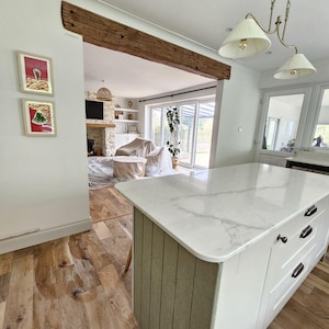 May include: A kitchen island with a white marble countertop and white cabinets. The island has a light green vertical paneling on the side. The floor is made of light brown wood planks. A wooden beam is above the opening to the living room.