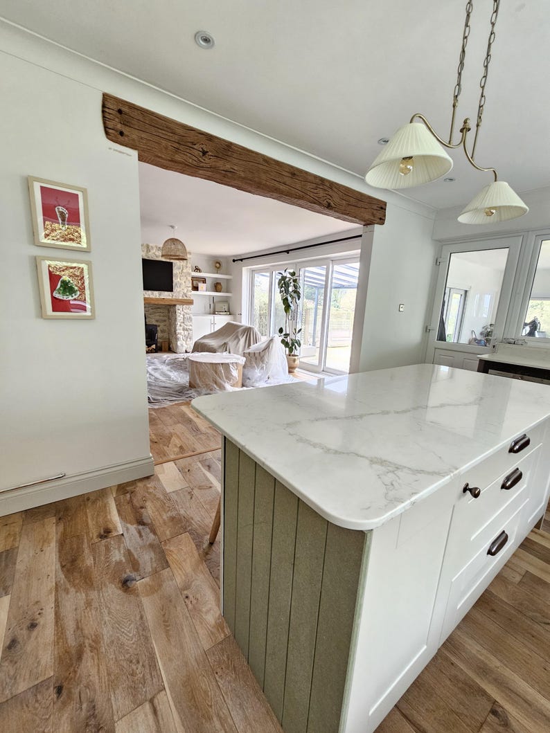 May include: A kitchen island with a white marble countertop and white cabinets. The island has a light green vertical paneling on the side. The floor is made of light brown wood planks. A wooden beam is above the opening to the living room.