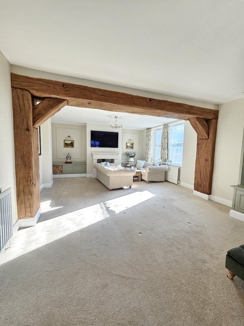 May include: A spacious living room with a neutral color palette. The focal point is a large, exposed wooden beam that frames the entrance. The room features a beige carpet, a white ceiling, and a fireplace with a mounted television.