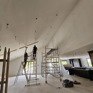 May include: Interior construction scene with two people working on a high, angled ceiling. Scaffolding and ladders are in use. The room is unfinished, with exposed concrete flooring and a large, open space.
