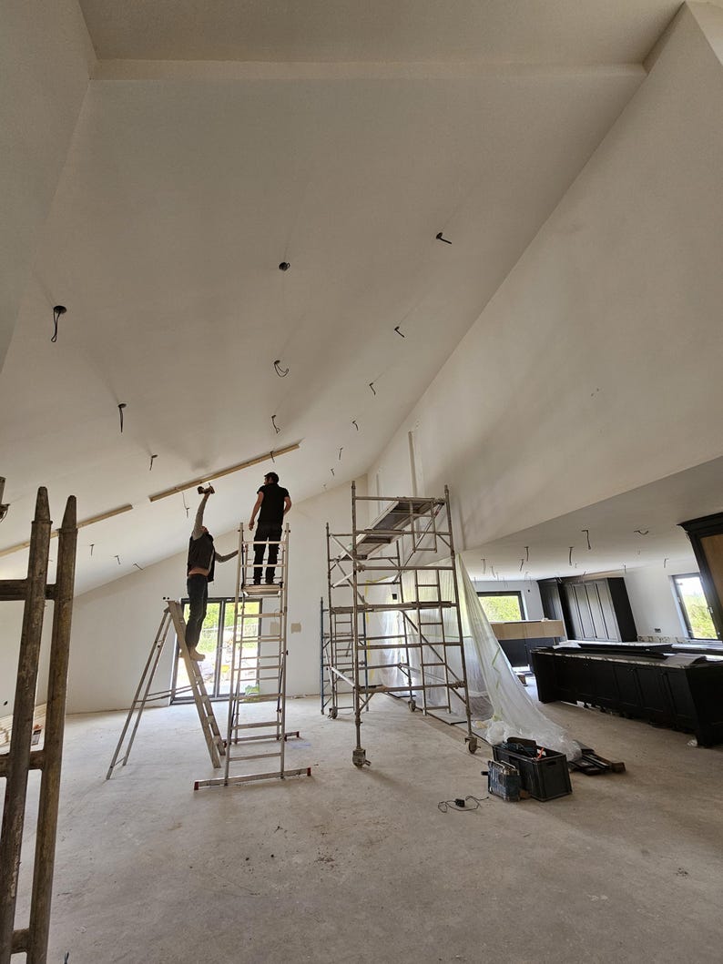 May include: Interior construction scene with two people working on a high, angled ceiling. Scaffolding and ladders are in use. The room is unfinished, with exposed concrete flooring and a large, open space.