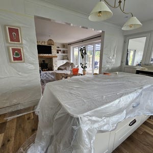 May include: A kitchen island covered in clear plastic sheeting, with white cabinets and black handles. The room is undergoing renovation, with walls and doorways also covered in plastic. A chandelier hangs above the island, and framed artwork is visible on the wall.
