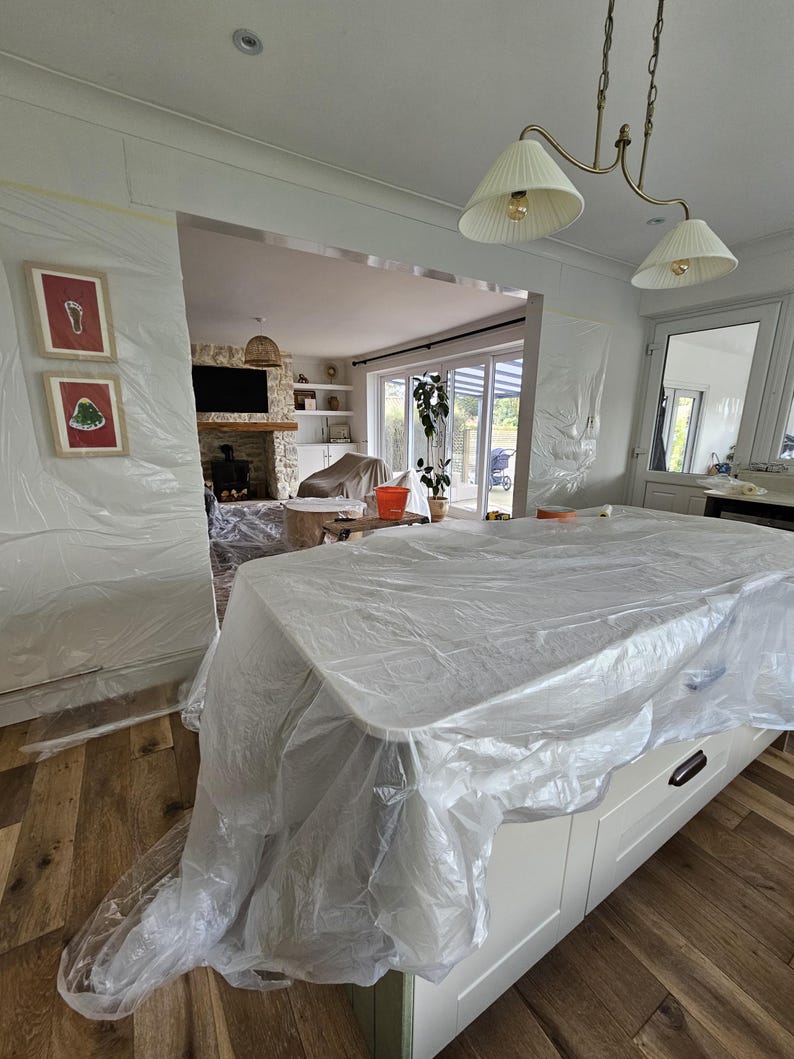 May include: A kitchen island covered in clear plastic sheeting, with white cabinets and black handles. The room is undergoing renovation, with walls and doorways also covered in plastic. A chandelier hangs above the island, and framed artwork is visible on the wall.