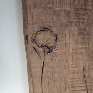 May include: Close-up of a weathered, brown wooden plank with a prominent knot. The wood grain is visible, with dark lines and textures. The plank has a natural, rustic appearance, with a rough, uneven edge.