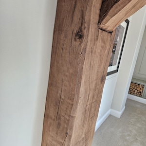 May include: Close-up of a weathered, brown wooden beam, part of a structural support. The wood grain is visible, with knots and natural imperfections. The beam is positioned against a white wall, with a glimpse of a framed picture and a carpeted floor.