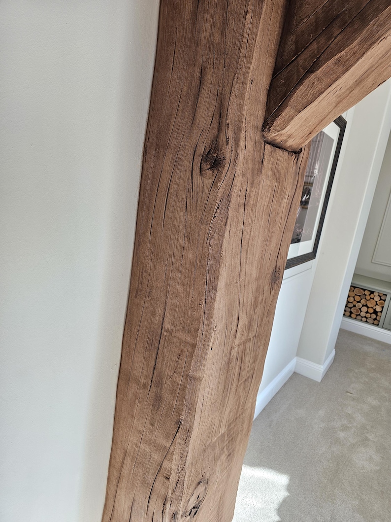 May include: Close-up of a weathered, brown wooden beam, part of a structural support. The wood grain is visible, with knots and natural imperfections. The beam is positioned against a white wall, with a glimpse of a framed picture and a carpeted floor.