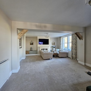 May include: An interior shot of a living room with neutral tones. The room features a gray carpet, beige walls, and a large opening leading to a seating area with a sofa and chairs. A fireplace and a television are visible.