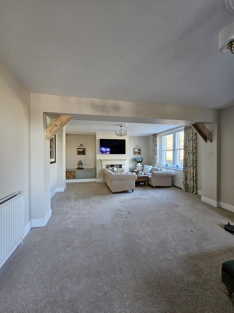 May include: An interior shot of a living room with neutral tones. The room features a gray carpet, beige walls, and a large opening leading to a seating area with a sofa and chairs. A fireplace and a television are visible.
