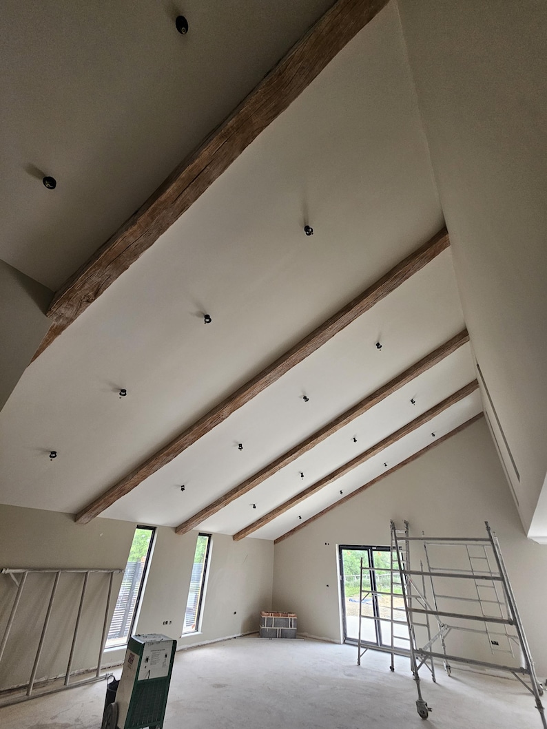 May include: Interior view of a room with a high, angled ceiling. The ceiling features exposed wooden beams and recessed lighting. The walls are painted a neutral color, and the room appears to be under construction, with a metal scaffolding visible.
