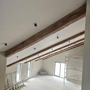 May include: Interior view of a room under construction, featuring a high, angled ceiling with exposed wooden beams. The beams are a warm brown color, contrasting with the white ceiling. Black light fixtures are installed. Windows and a door are visible.