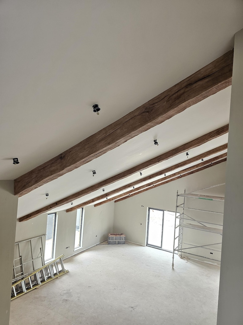 May include: Interior view of a room under construction, featuring a high, angled ceiling with exposed wooden beams. The beams are a warm brown color, contrasting with the white ceiling. Black light fixtures are installed. Windows and a door are visible.